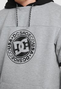 Felpa grigia con cappuccio e logo nero che presenta le lettere "DC" al centro. Include un cappuccio nero e lacci con dettagli bianchi.