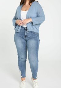 Jasnoniebieski dziergany kardigan na białej bluzce, w połączeniu z niebieskimi dżinsami ozdobionymi białą koronką na dole oraz białymi tenisówkami.