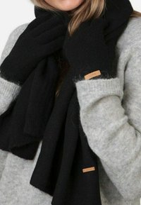 Gants et écharpe en tricot noirs, fabriqués en matériau doux, enroulés autour du cou. L'étiquette de l'écharpe présente un accent marron clair. Un pull gris est visible.