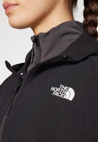 Femeie îmbrăcată cu o jachetă neagră North Face cu fermoar, suprapusă peste un top gri închis cu guler înalt, arătându-i partea inferioară a feței și părul împletit.