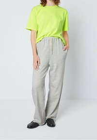 Person som bär en ljusgrön kortärmad skjorta instoppad i gråa dragbandsbyxor, med en hand i fickan och svarta slipskor.