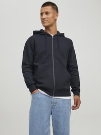 Navy-blaues Zip-Hoodie aus weichem Material mit Kängurutasche, verstellbarem Kordelzug an der Kapuze, gerippten Bündchen, getragen über einem weißen Shirt und hellblauen Jeans.