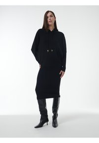 Schwarzes Strick-Hoodie-Kleid mit Kordelzügen, gerippter Textur und lockerer Passform. Kombiniert mit schwarzen kniehohen Stiletto-Stiefeln.
