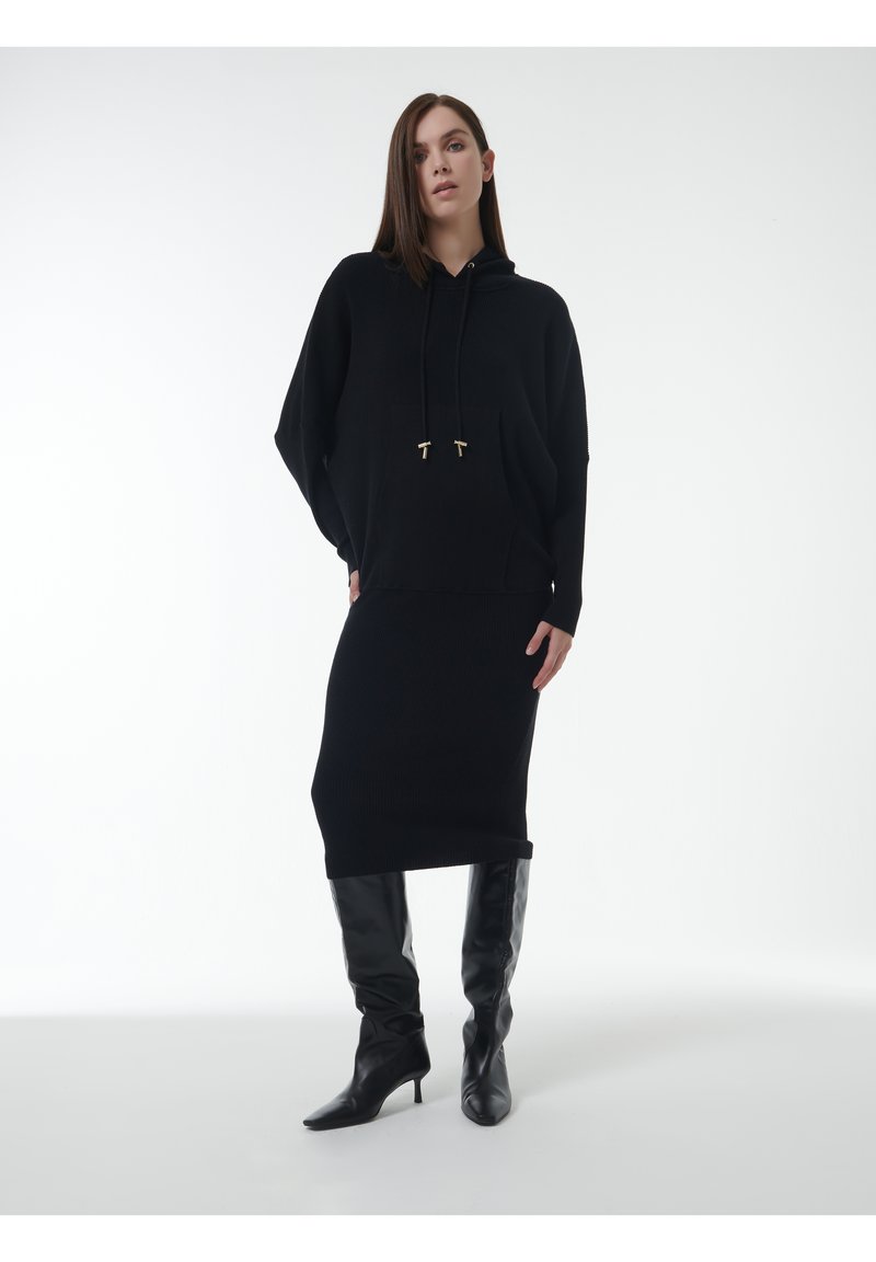 Schwarzes Strick-Hoodie-Kleid mit Kordelzügen, gerippter Textur und lockerer Passform. Kombiniert mit schwarzen kniehohen Stiletto-Stiefeln.