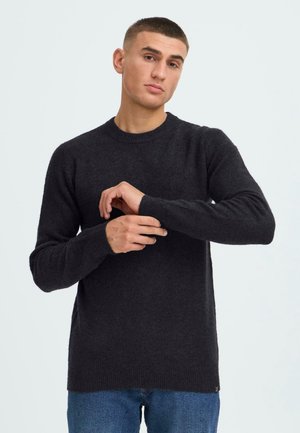 Ung mann som justerer ermet på en svart strikket genser, står mot en ensfarget lys bakgrunn, iført blå jeans.