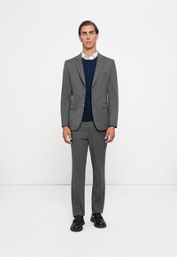 Grauer maßgeschneiderter Anzug mit zwei Knöpfen, darunter ein navyfarbener Pullover und ein weißes Hemd mit Kragen. Schwarze Schuhe runden das Outfit ab.