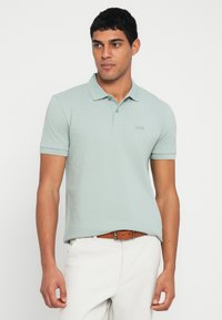 Svetlozelené polokošeľa s golierom a krátkymi rukávmi. Má jemné logo na hrudi a je vyrobená z pletenej látky. Nosená s béžovými nohavicami.