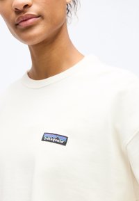 Κοντινό πλάνο ενός ατόμου που φοράει λευκό πουκάμισο Patagonia, με το λογότυπο στο στήθος να φαίνεται σε απλό φωτεινό φόντο.