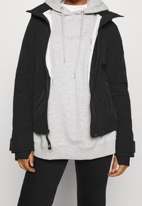 Svart ytterjacka med en figursydd design och varmt tyg, lager-på-lager över en ljusgrå hoodie med dragsko. Modellen bär svarta jeans.