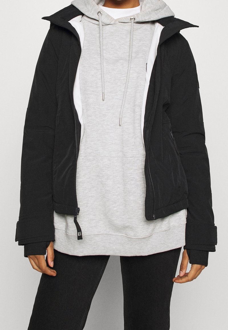 Svart ytterjacka med en figursydd design och varmt tyg, lager-på-lager över en ljusgrå hoodie med dragsko. Modellen bär svarta jeans.