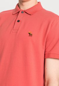 Punainen polo-paita, joka on valmistettu puuvillasekoitteesta. Siinä on kolme nappia edessä ja brodeerattu hirvimerkki vasemmassa rinnassa. Lyhyet hihat.