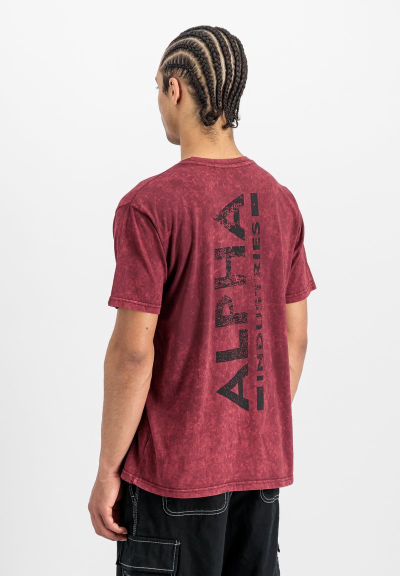 Červené tričko s texturou a vybledlým vzhledem. Na zadní straně má velký černý potisk "ALPHA INDUSTRIES". Krátké rukávy, volný střih.