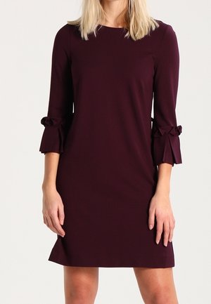 Femme portant une robe violet foncé, longue jusqu'aux genoux, à manches longues avec des manches évasées et des détails de nœud aux coudes.