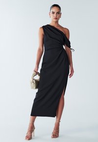 Juoda vienos peties suknelė iš lygaus audinio, su sukryžiuota detale juosmenyje ir aukšta skeltimi. Derinama su auksinėmis basutėmis su dirželiais ir pintine rankine.