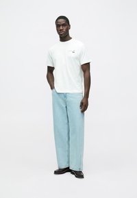 Biały bawełniany T-shirt z krótkimi rękawami i małym logo, w połączeniu z jasnoniebieskimi szerokimi dżinsami i czarnymi butami, stojący na neutralnym tle.