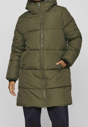 Person iført lang olivengrøn polstret vinterfrakke med frontlynlås og sidelommer, med en hånd i lommen, synlig fra skuldrene til midt på låret.
