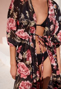 Blommig överdel med puffärmar, med svart bas och stora rosa rosor. Knuten i midjan med tunna band, lager-på-lager över en svart bikini.