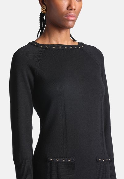 Femme portant un pull en maille noir à manches longues avec de petits détails dorés sur l'encolure et les poches, ainsi que de grandes boucles d'oreilles rondes dorées.
