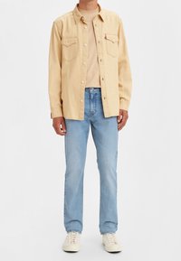 Camicia beige con bottoni e due tasche sul petto, indossata sopra una camicia beige, abbinata a jeans azzurri chiari e sneakers crema. Tessuto dalla consistenza liscia.