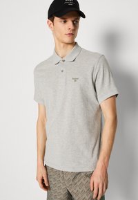 Ανοιχτό γκρι πόλο μπλουζάκι από μαλακό βαμβάκι, με μια διακριτική πράσινη κεντημένη λογότυπο και δύο κουμπιά στην πλαϊνή πλευρά. Συνδυασμένο με μοτίβο σορτς.