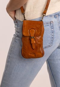 Bolso cruzado de cuero marrón con solapa frontal, correa ajustable y detalles de costura. Diseño compacto, complementado por un logo de marca.