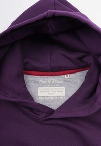 Lila bomullshoodie med en mjuk insida. Har en grå etikett som anger 100% ekologiskt material och en röd detalj vid halsringningen.