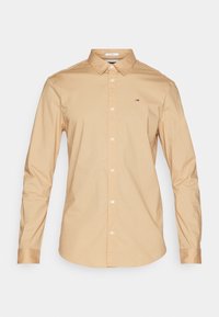 Camisa de manga larga en un suave tejido de algodón color beige claro, con cuello abotonado y cierre de botones en la parte delantera. Presenta un pequeño logo en el pecho.