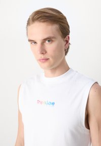 Camisa sin mangas blanca de algodón con la inscripción "Esto es Amor" en colores del arcoíris en degradado. Textura suave con cuello redondo.