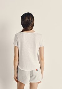 Sieviete ar gariem brūniem matiem, sasietiem zemu zirgastē, valkājot baltu īsroku krekls un gaiši pelēkas šortus, stāvot ar muguru pretī.