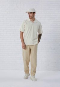 Hellbeiges Poloshirt mit kurzen Ärmeln, einzelne Brusttasche, kombiniert mit beigen Hosen und weißen Turnschuhen, vor einer strukturierten weißen Wand.