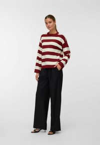 Pull rayé bordeaux et crème avec une coupe ample, manches longues, col côtelé, associé à un pantalon noir à taille haute et jambes larges.