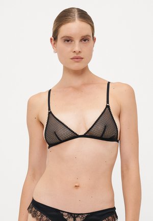 Mujer de piel clara con el cabello peinado hacia atrás, vistiendo un sujetador de encaje negro transparente y ropa interior a juego, contra un fondo blanco liso.