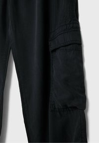 Gros plan sur un pantalon noir montrant une grande poche cargo latérale avec rabat et coutures apparentes sur un tissu lisse.