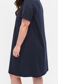 Osoba w granatowej sukience do kolan z krótkimi rękawami i białymi groszkami, przedstawiona od połowy ramienia do kolan na jednolitym tle.