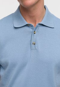 Camisa polo de color azul claro con una textura de punto, que cuenta con un cuello y tres botones en el cuello. Los botones son de color beige.