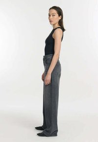 Zwarte mouwloze top gecombineerd met hoog-gesneden, loszittende grijze jeans. Model staat zijwaarts, waardoor de textuur en het ontwerp van de spijkerbroek goed zichtbaar zijn.