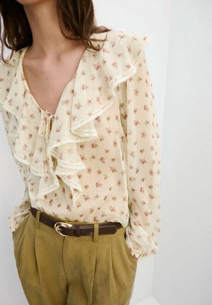 Mujer con una blusa cremosa transparente con pequeñas flores rosas y cuello con volantes, metida dentro de pantalones caquis de talle alto con un cinturón oscuro.