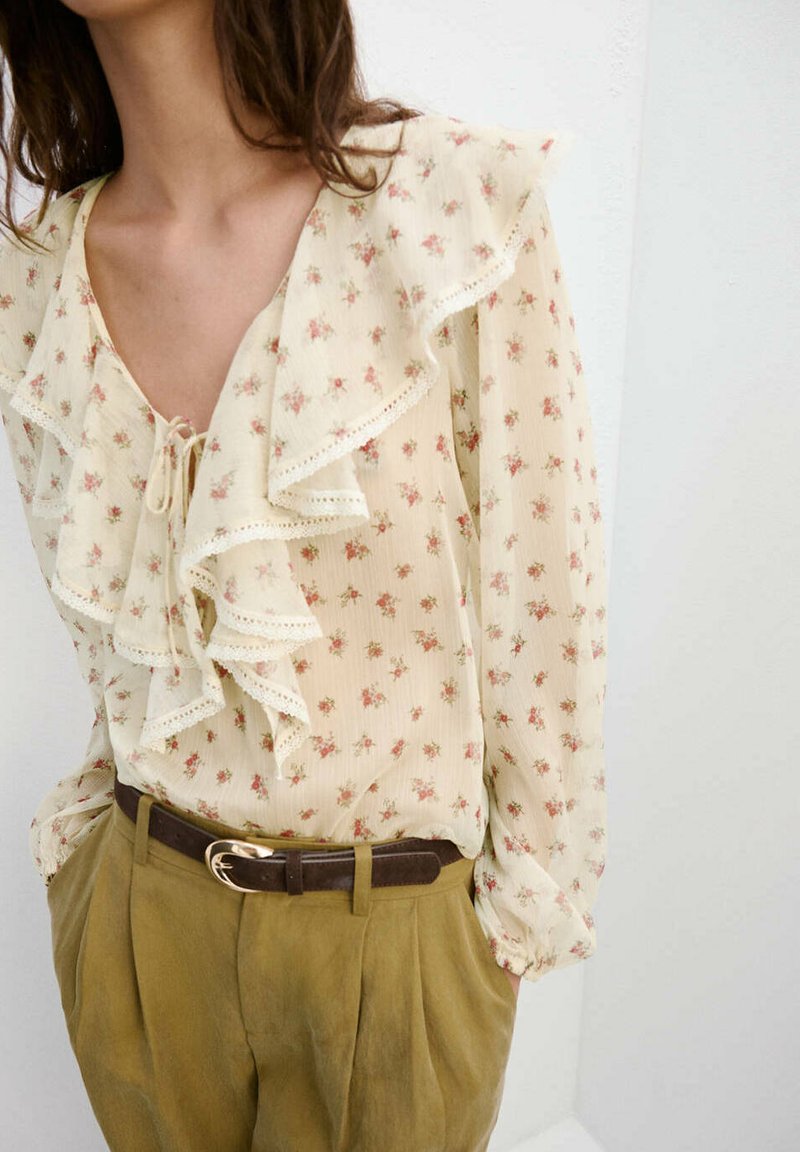 Femme portant un chemisier crème transparent avec de petites fleurs roses et un col volanté, rentré dans un pantalon taille haute kaki avec une ceinture foncée.