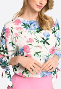 Bluse mit Blumenmuster in Creme mit rosa und blauen Blumen. Hat lange Ärmel mit gebundenen Manschetten. Glatte Stoffqualität und lässiger Schnitt.