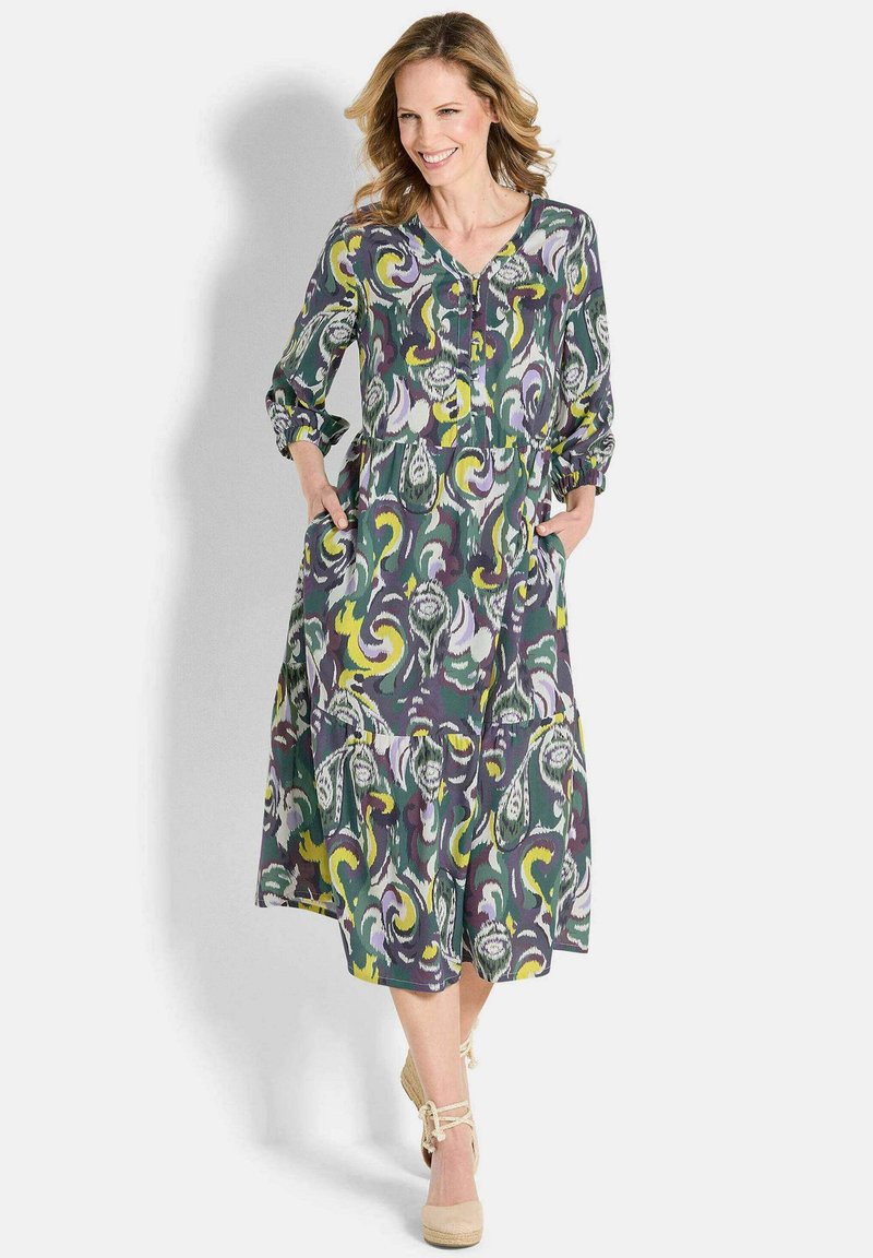 Frau, die ein farbenfrohes, gemustertes Midi-Kleid mit Dreiviertelärmeln und Taschen trägt, lächelnd und nach vorne gehend in beigen Keilsandaletten.