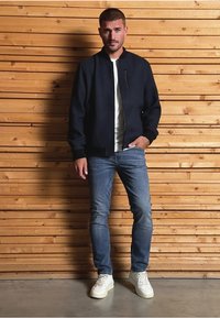 Chaqueta de bombardero navy con ribetes negros, camiseta blanca, vaqueros azul claro y zapatillas blancas. El modelo está de pie contra una pared de madera.