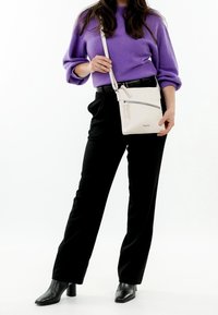 Femme portant un pull violet et un pantalon noir, tenant un sac bandoulière blanc avec une poche avant zippée.