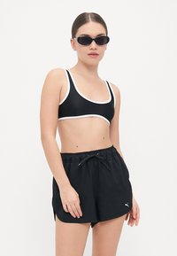 Črn atletski bikini zgornji del z belim obrobom in ujemajočimi črnimi kratkimi hlačami z vrvico. Model nosi temna sončna očala.