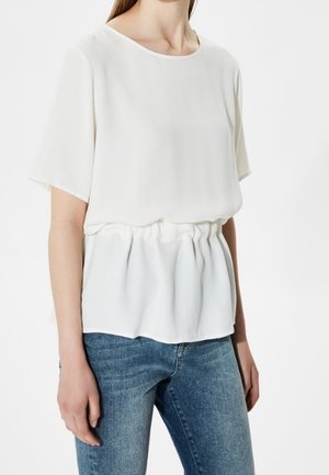 Woman wearing a loose white short-sleeve blouse with a gathered waist paired with blue denim jeans on a white background.
