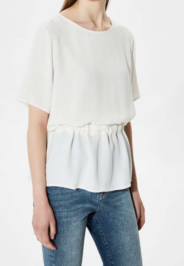 Femme portant un chemisier blanc ample à manches courtes avec une taille froncée, associé à un jean en denim bleu sur fond blanc.