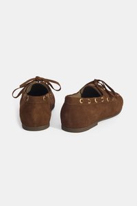 Braune Wildleder-Slipper mit Schnürdetails, goldenen Ösen und flacher Sohle, mit abgerundeter Spitze und schlichter, klarer Designsprache.