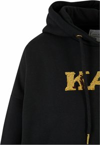 Čierna mikina s kapucňou, zdobená zlatým flitrovým nápisom. Materiál sa zdá byť mäkký a má textúrovaný povrch s detalovaním na šnúrkach.