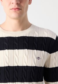 Megzta megrišė, turinti pakaitinius tamsiai mėlynus ir kreminius horizontalius dryžius, iškloto pynimo tekstūrą ir mažą emblema ant krūtinės.