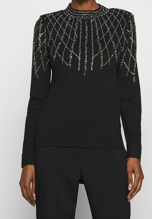 Haut noir à manches longues avec un design en sequins argentés en forme de toile sur les épaules et la poitrine, porté avec un pantalon noir, modèle se tenant debout avec les mains détendues.