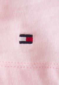 Tessuto di cotone rosa con un piccolo logo ricamato che presenta strisce orizzontali nere, bianche e rosse vicino al fondo. Dettaglio dell'orlo cucito visibile.